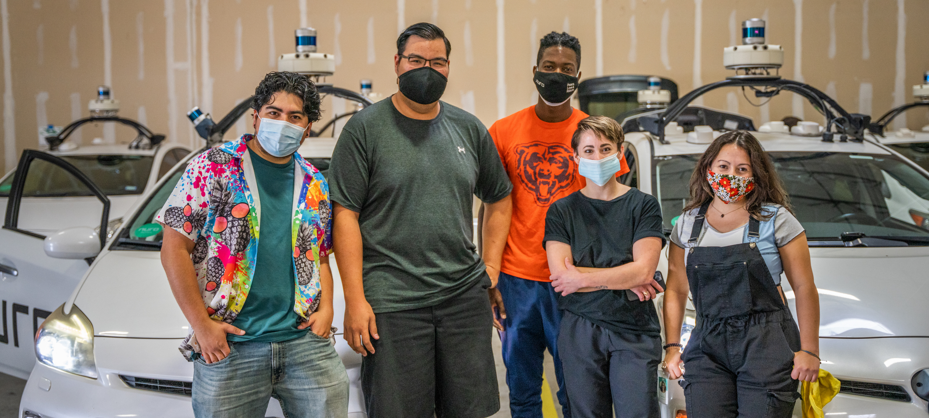 Group of diverse Nuro employees wearing face masks and standing next to Nuro vehicles