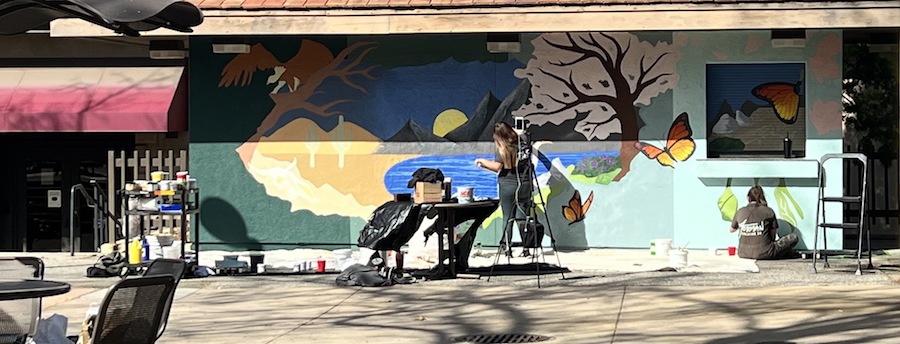 two students, one on ladder and one sitting on ground, painting mural outside Campus Center
