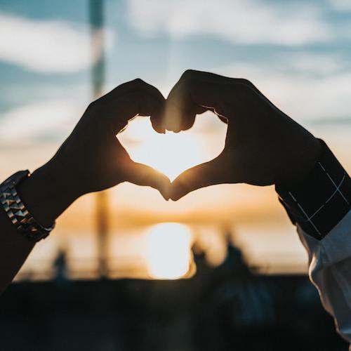 two hands making a heart silhouetted against sunrise