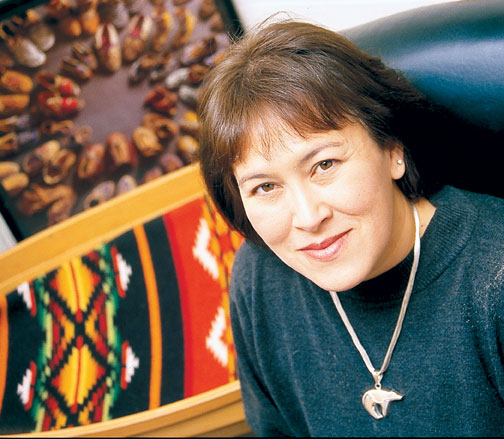 Lori Arviso Alvord MD has a closed mouth smile. She has short brown hair and is wearing a bear necklace with a blue greenish sweater. There is Native Amerian art in the background.. 