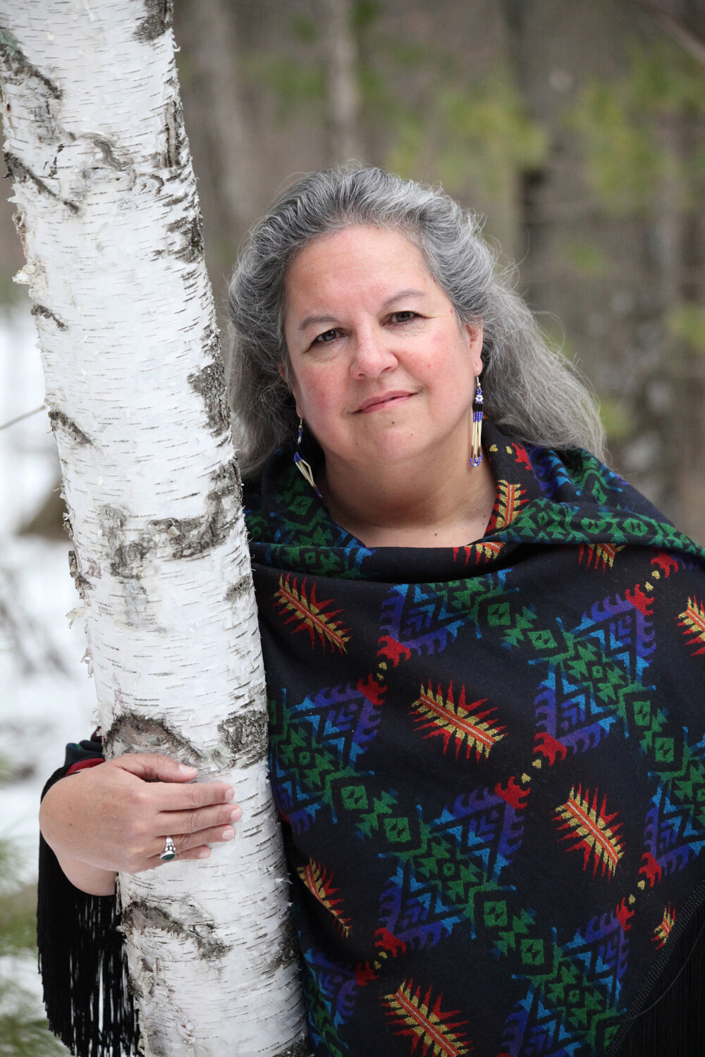 Robin Wall Kimmerer has her arm around a birch tree. Her hair is salt and pepper colored. She is wearing a Native American pattern shawl with dangling earrings made of beads and bone.